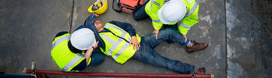 Scène d’accident sur un chantier. Deux personnes portent secours à un homme allongé à terre. Les trois individus portent un gilet jaune à bandes réfléchissantes